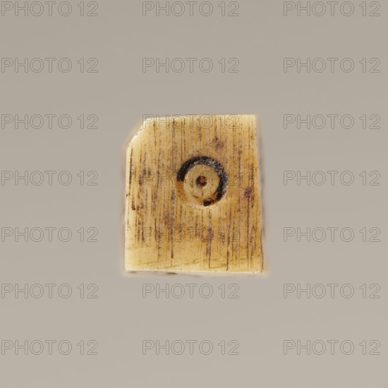 Game die made of animal bone with six unequal sides. Each side is engraved with a number from 1 to 6 as circles with a dot in the center. 4th century, Egypt. Height 0.7 cm, width 0.7 cm, depth 0.7 cm., A small rectangular object with slightly rounded corners sits against a plain beige-gray background; the object is primarily yellowish-beige with vertical fine brown striations and darker brown spots and smudges, and it has a circular depression near its center containing concentric rings of darker brown and black tones.