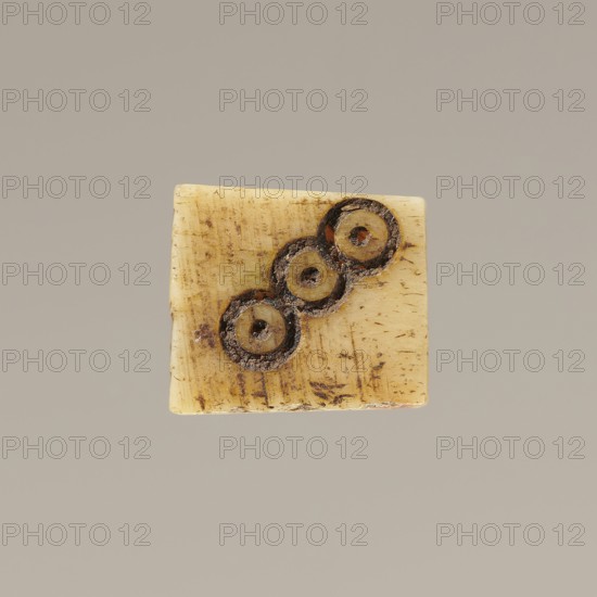Game die made of animal bone with six unequal sides. Each side is engraved with a number from 1 to 6 as circles with a central dot. 4th century, Egypt. Height 0.7 cm, width 0.7 cm, depth 0.7 cm., A small square object with a smooth, slightly worn surface in a pale yellowish-cream color sits against a neutral light gray background; the square shows a few linear scratches and subtle brownish discolorations, and contains a diagonal arrangement of a few circular depressions or rings that are darker brown and show textured, rough edges with crumbly dark material inside, creating contrast with the lighter surface.