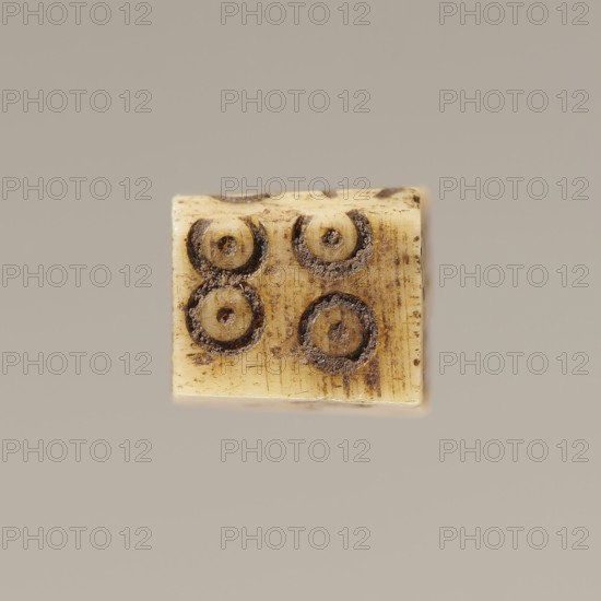 Game die made of animal bone with six unequal sides. Each side is engraved with a number from 1 to 6 in the form of circles with a dot in the center. 4th century, Egypt. Height 0.7 cm, width 0.7 cm, depth 0.7 cm., A small rectangular object with a worn, creamy beige surface sits against a plain gray-beige background; the object shows a few circular impressions with dark brown residue and rough, textured edges, and there are subtle vertical striations and darker brown smudges across its face.