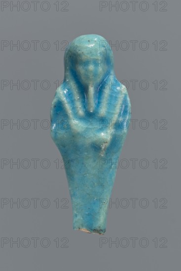 Shabti, mummy-shaped figurine, funerary gift. Back supported by a pillar/pilaster. Legs missing up to knee height. Arms crossed on the chest, right folded over left. Right hand holds a hoe; left hand holds a pickaxe. Wearing a three-part smooth wig. Long, straight, braided beard. Facial details poorly visible. Not inscribed. 664–332 BC Late Period, Egypt. Height 6.9 cm., A small carved figure rendered in a glossy turquoise-blue material against a plain gray background, showing a stylized human form with a rounded head, a simplified face with faint features, broad shoulders that narrow into a tapered body, and a raised collar or headdress shape framing the face; the surface displays subtle variations of blue and greenish-blue tones, lighter highlights where the gloss catches light, and small darker specks and tiny surface cracks, with a slightly rougher chipped edge at the bottom.