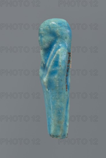 Shabti, mummy-shaped figurine, funerary gift; back supported by a pillar/pilaster. Legs missing up to knee height. Arms crossed on the chest, right folded over left. In the right hand is a hoe, in the left a pickaxe. On the head is a three-part, smooth wig. The beard is long, straight, and braided. Facial details are poorly visible. Not inscribed. 664–332 BC, Late Period, Egypt. Height 6.9 cm., A small carved object shaped like a standing figure with a rounded head and draped body, rendered in a smooth blue-green material with variations of turquoise and aquamarine tones, showing areas of lighter and darker blue-green, some pale greenish tints, and tiny dark specks and weathered spots; the surface has a slightly glossy, worn appearance with subtle ridges and folds suggesting drapery, and the object is photographed against a plain gray background.