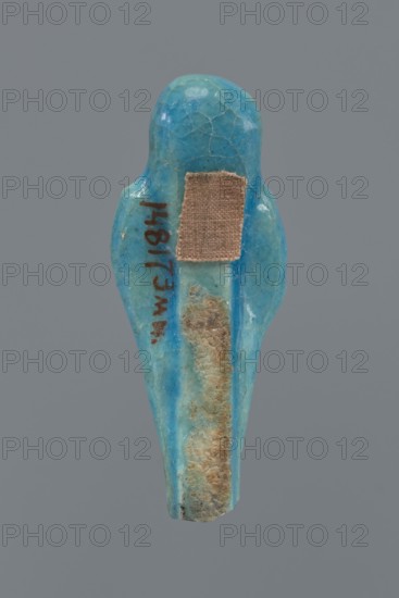 Shabti, mummy-shaped figurine, funerary gift. Mummy-shaped figurine with back supported by a pillar/pilaster. Legs missing up to knee height. Arms crossed on the chest, right folded over left. Right hand holds a hoe; left hand holds a pickaxe. Wearing a three-part smooth wig. Long, straight, braided beard. Facial details poorly visible. Not inscribed. 664–332 BC, Late Period, Egypt. Height 6.9 cm., A small carved object shaped like a stylized bird with a rounded head and tapered body sits against a plain gray background, showing a smooth glossy surface in shades of bright turquoise and teal with areas of lighter blue and pale green; a rectangular patch of beige-brown fabric is adhered near the upper back, and a vertical strip of rough tan-brown encrustation runs along the center of the lower body, while a few thin dark brown marks appear on one side.