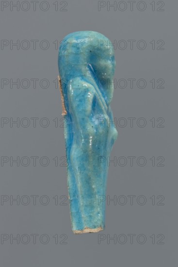 Shabti, Mummy-shaped figurine, back supported by a pillar/pilaster. Legs missing up to knee height. Arms crossed on the chest, right folded over left. In the right hand is a hoe, in the left a pickaxe. On the head is a three-part, smooth wig. The beard is long, straight, and braided. Facial details are poorly visible. Not inscribed. 664–332 BC, Late Period, Egypt. Height 6.9 cm., A small sculpted figurine shaped like a standing person shown in profile, rendered in a bright turquoise blue with lighter and darker blue variations and a few tiny dark specks, with a slightly glossy surface and visible cracks and wear; the figure has a rounded head, a subtle suggestion of a shoulder and arm, and a tapered body ending in a rough beige edge, all set against a plain medium-gray background.