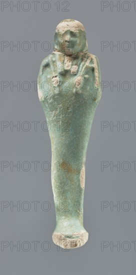 Shabti, Mummy-shaped figurine, funerary gift, 664–332 BC Late Period, Egypt. Standing on a base, back supported by a pillar/pilaster. Arms crossed on the chest with only the hands protruding from under the shroud; right hand holds a hoe, left hand a pickaxe, both tool ends wrapped. Basket on the back slung on a single twisted cord over the left shoulder. Wears a three-part smooth wig that merges into the pillar, with a long braided beard. Facial details poorly visible. Not inscribed. Height 12.5 cm., A single elongated figurine with a rounded top resembling a head and a tapered lower body, shown against a solid neutral background; the surface of the object is predominantly turquoise with areas of lighter greenish-blue and pale beige where the outer layer is worn; the top has a beige patch and the face area shows subtle raised features with a mix of turquoise and beige; the shoulders and upper torso reveal faint raised details and small worn spots of beige; the midsection displays a tex...