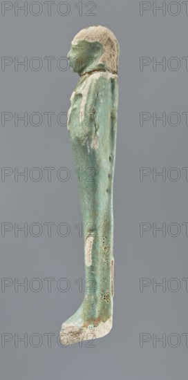 Shabti, mummy-shaped figurine, funerary gift; standing on a base with back supported by a pillar/pilaster. Arms crossed on the chest with only the hands protruding from under the shroud. Right hand holds a hoe and left hand a pickaxe, both tool ends wrapped. A basket is on the back, slung on a single twisted cord over the left shoulder. Wears a three-part smooth wig that merges into the pillar; long braided beard. Facial details poorly visible. Not inscribed. 664–332 BC, Late Period, Egypt. Height 12.5 cm., A narrow vertical statuette of a standing human figure photographed in profile against a plain gray background, with a rounded head and simplified facial features, a long wrapped body forming a straight column, and visible surface wear and erosion; the object shows a predominant turquoise-green color with patches of off-white and beige where the surface is worn, and small dark pitted areas on the side.