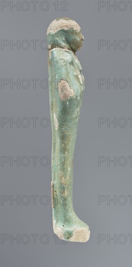 Shabti, mummy-shaped figurine, funerary gift, hoes; standing on a base, back supported by a pillar/pilaster. Arms crossed on the chest with only the hands protruding from under the shroud. Right hand holds a hoe, left a pickaxe; the ends of both tool handles are wrapped. A basket is on the back, slung on a single twisted string over the left shoulder. Wears a three-part smooth wig that merges into the pillar and a long braided beard. Facial details are poorly visible. Not inscribed. 664–332 BC, Late Period, Egypt. Height 12.5 cm., A narrow vertical turquoise-green object with areas of pale beige and off-white erosion, shaped like a standing human figure viewed in profile, showing a rounded head, a draped upper body, an elongated straight lower body and a flat foot; the surface has a weathered, pitted texture with several rough patches and small holes, and the background is a uniform cool gray.