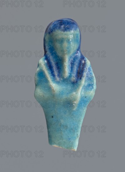 Shabti, Mummy-shaped figurine used as a funerary offering, supported at the back on a pillar. Legs missing from the knees down. Arms crossed on the chest, right folded over left. In the right hand is a mattock, in the left an axe. On the back is a bag slung on a string over the left shoulder. Convex tools. On the head is a smooth, tripartite wig and a long, straight beard. Not inscribed. 664–332 BC, Late Period, Egypt. Shabti; height 6 cm., A small figurine-shaped object with smooth, rounded contours in shades of blue and turquoise, showing a darker blue area on the top and along the sides of the head and shoulders, with lighter turquoise on the face, chest, and lower portion; the surface shows a slightly glossy, worn texture with visible fine cracks and a few small darker specks, all set against a plain gray background.