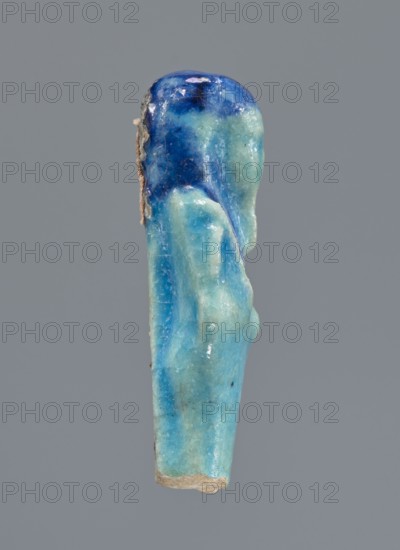 Shabti, Mummy-shaped figurine supported at the back on a pillar. Legs missing from the knees down. Arms crossed on the chest, right folded over left. In the right hand is a mattock, in the left an axe. On the back is a bag slung on a string over the left shoulder. Convex tools. On the head is a smooth, tripartite wig. Long, straight beard. Not inscribed. 664–332 BC; Late Period, Egypt. Shabti, height 6 cm., A vertical small sculpted object shown against a plain gray background, predominantly in shades of blue and turquoise with patches of darker blue near the top and lighter turquoise toward the lower portion; the surface appears glossy and slightly irregular with some small areas of rough beige material along one side and at the base, and subtle variations in texture and faint surface scratches are visible.