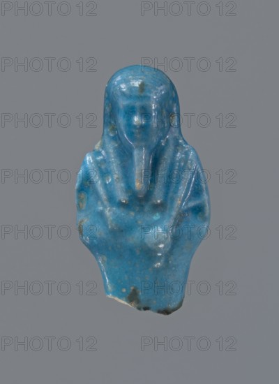 Shabti, Mummy-shaped figurine, funerary offering, supported on a pillar at the back. Legs missing. Arms crossed on the chest, right folded over left. In the right hand is a hoe, in the left a pickaxe. On the back is a bag slung on a string over the left shoulder. Convex tools. On the head is a long, smooth, tripartite wig. Long, straight beard braided. Not inscribed. 664–332 BC Late Period, Egypt. Shabti, height 5.2 cm., A small sculpted figurine of a human-like figure shown against a plain gray background, rendered in a vibrant blue with areas of lighter blue and small patches of darker blue and greenish discoloration, the figure has a rounded head with simplified facial features and a narrow beard-like projection, shoulders and torso suggested with smoothed, flowing contours and a few subtle ridges, surface appears glossy and slightly pitted with some worn spots and a chipped lower edge revealing a lighter interior.
