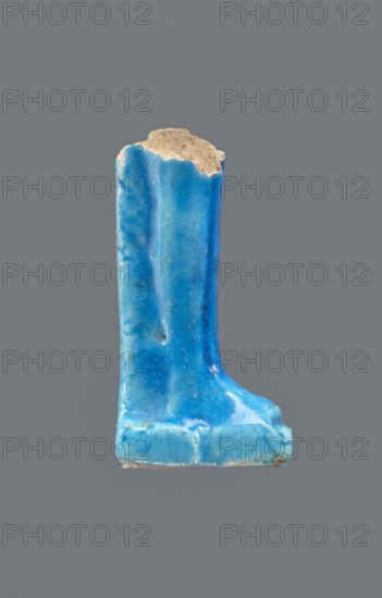 Shabti legs, Fragment of the legs of a mummy-shaped shabti figurine preserved from the knees down. The feet rest on a base, the back supported by a pilaster. On the front of the legs is a painted hieroglyphic inscription in vertical lines. Late Period, Egypt, 664–332 BC. Material: shabti. Height 8.5 cm; width 2 cm., A single ceramic fragment shaped like a short, upright boot or column with a flat base, glazed in several shades of blue including bright sky blue, deeper cobalt blue, and a few turquoise tones, with a small area of exposed beige ceramic at the top; the glaze surface shows glossiness, subtle streaks and mottling, and a few small surface pits and rough edges, all set against a plain gray background.