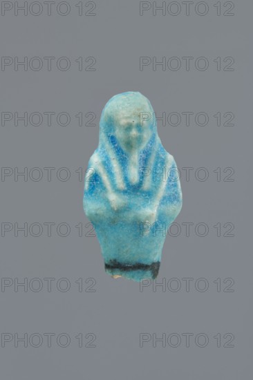 Shabti, Mummy-shaped figurine, funerary gift, supported on a pillar at the back. Legs missing. Arms crossed on the chest, right folded over left. In the right hand, a hoe; in the left, a pickaxe. On the back, a bag slung on a string over the left shoulder. Convex tools. On the head, a long, smooth, three-part wig. Long, straight beard braided in a plait. Facial details poorly visible. Not inscribed. 664–332 BC Late Period, Egypt. Height 5.4 cm., A small carved figurine in shades of turquoise and light blue with darker blue accents, showing a simplified humanlike form with a hooded head and folded arms, some pale greenish-blue areas and tiny brownish specks on the surface, and a narrow dark band near the base, all set against a solid medium-gray background.
