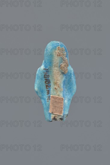 Shabti, Mummy-shaped figurine, funerary gift, supported on a pillar at the back. Legs missing. Arms crossed on the chest, right folded over left. In the right hand a hoe, in the left a pickaxe. On the back a bag slung on a string over the left shoulder. Convex tools. On the head a long, smooth, three-part wig. Long, straight beard braided. Facial details poorly visible. Not inscribed. 664–332 BC, Late Period, Egypt. Height 5.4 cm., A small blue object with an irregular rounded top and slightly flared lower sides sits against a solid gray background; its surface shows areas of lighter blue and patches of beige and brown encrustation, and a small square patch of tan fabric is attached near the lower portion, with some darker grayish spots along the bottom edge.