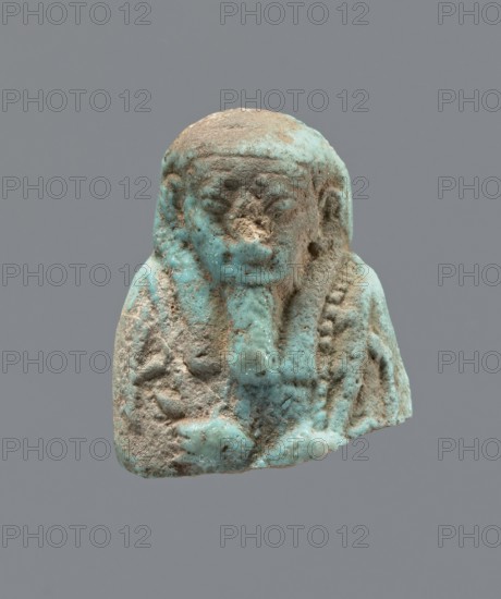 Ushabti - upper part of a figurine. Mummy-shaped funerary figurine ushabti, head and torso fragment. Figurine resting on a pillar with arms crossed on the chest, hoes in both hands. A twisted rope runs from the right hand over the left shoulder to the back. Convex tools; a basket hangs on the back on a string; the basket is engraved. Three-part smooth wig, long wide smooth beard. Large almond-shaped eyes, large ears; other facial details damaged. Inscription placed in a column on the pillar, text illegible. 664–332 BC, Late Period, Egypt. Height 3.5 cm., A small sculpted bust with a rough, weathered surface showing a mix of turquoise blue and sandy beige colors, depicting a rounded head with a stylized headdress or hair framing the face, a worn facial area with shallow eye and mouth forms, a vertical element at the front resembling a braided or folded feature, textured areas on the shoulders and chest with traces of turquoise glaze, and small patches of exposed beige material where the...
