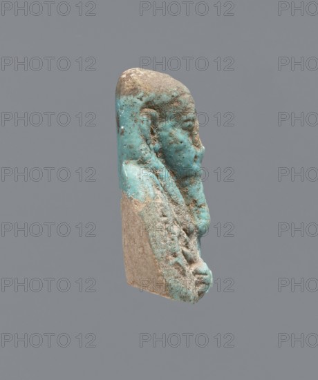 Ushabti - upper part of a figurine. Mummy-shaped figurine, funerary gift; head and torso fragment. Figurine resting on a pillar; arms crossed on the chest holding hoes in both hands. In the right hand, a twisted rope runs over the left shoulder to the back. Convex tools. A basket hangs on the back on a string; basket is engraved. On the head is a three-part smooth wig; long, wide, smooth beard. Large almond-shaped eyes, large ears; other facial details damaged. Inscription placed in a column on the pillar text illegible. 664–332 BC, Late Period, Egypt. Ushabti; height 3.5 cm., A small sculpted fragment showing the profile of a human head and upper torso, with a weathered surface and rough texture; the piece displays a predominant turquoise blue glaze or pigment on much of the visible surface, along with areas of beige and light brown where the underlying material is exposed and worn; the facial features including a nose, lips, and a recessed eye are visible in profile, and a headdress ...