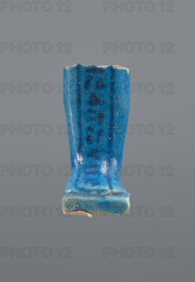 Shabti legs, mummy-shaped figurine fragment: fragment of the legs of a mummy-shaped shabti figurine preserved from the knees down. The feet rest on a base, the back of the figurine is supported by a pilaster. In front of the legs is a column of hieroglyphic inscription enclosed in a frame; painted inscription. Late Period, Egypt, 664–332 BC. Shabti; height 4.3 cm, width 2 cm., A blue glazed ceramic or faience fragment shaped like a small upright rectangular column with a flared top and a stepped base sits against a plain gray background, showing worn edges and small chips where an underlying beige material is visible; the glaze displays variations of bright turquoise blue, deeper cobalt blue streaks, and some darker blue spots, with a few areas of lighter bluish-green near the base and tiny specks of pale beige from surface wear.