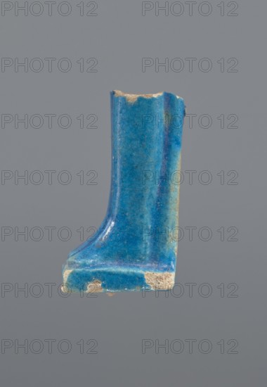 Shabti legs, mummy-shaped figurine fragment: fragment of the legs of a mummy-shaped shabti figurine preserved from the knees down. The feet rest on a base, the back of the figurine is supported by a pilaster. In front of the legs is a column of hieroglyphic inscription enclosed in a frame. Painted inscription. Late Period, Egypt, 664–332 BC. Shabti. Height 4.3 cm, width 2 cm., A fragment of an object with a glossy blue surface showing variations of bright blue and turquoise, with areas where the blue glaze is chipped revealing a beige, sandy inner material; the fragment has a vertical, slightly curved face rising from a flat, rectangular base, and the edges show irregular breaks and small rough spots against a plain gray background.
