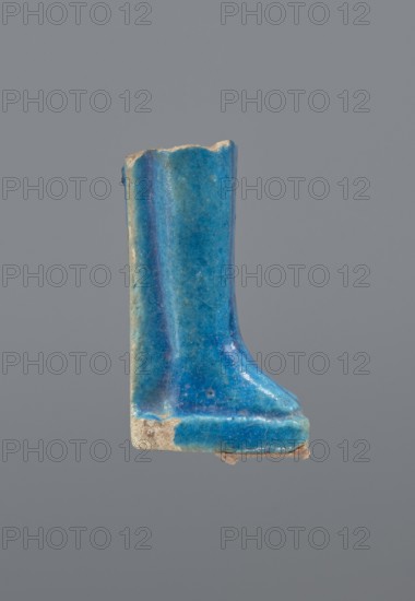 Shabti legs, mummy-shaped figurine, funerary gift. Fragment of the legs preserved from the knees down; the feet rest on a base and the back is supported by a pilaster. In front of the legs is a column of hieroglyphic inscription enclosed in a frame; painted inscription. Late Period, Egypt, 664–332 BC. Shabti; height 4.3 cm, width 2 cm., A glazed ceramic fragment shaped like a stylized boot or shoe, shown against a plain gray background, with a glossy surface in shades of blue including turquoise and deeper blue, and areas of lighter blue-green; the glaze shows speckled texture and small worn spots revealing a beige or sandy clay underneath, and there are rough, unglazed beige edges at the top and bottom.