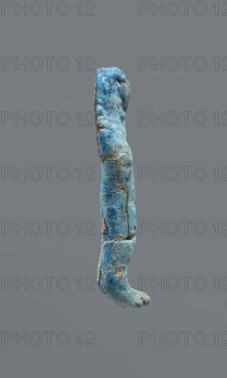 Shabti, Mummy-shaped figurine, funerary gift. Legs missing. Arms crossed on the chest; hands invisible under the shroud. Smooth, tripartite wig on the head. Facial details invisible. Not inscribed. 664–332 BC; Late Period, Egypt. Height 3.7 cm., A single elongated object suspended against a plain gray background, showing a weathered, irregular surface with a dominant blue color and areas of lighter and darker blue, small patches of red along one side, and touches of gray and sandy beige where material appears chipped or worn; the object has a curved, uneven profile with rough textures, pitting, and small granular deposits visible across its surface.