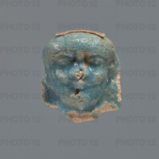 Shabti head, mummy-shaped figurine, funerary gift. Head of a shabti figurine depicting a king: oval face with full cheeks; prominent eyebrow arch; eye and nose shapes difficult to distinguish; corners of the mouth slightly upturned. Nemes placed low on the forehead, edges slightly outlined; end of the rear pillar visible at back of head. Ptolemaic Period, 332–30 BC, Egypt. Shabti; height 3 cm, width 2.3 cm., A small sculpted head fragment with a glossy turquoise-blue surface and rough beige areas where material is missing, showing the contours of a forehead, eye sockets, a nose area with chipped sections, and a partial mouth, with a flat beige strip across the top and uneven beige edges along the sides and bottom against a plain gray background.
