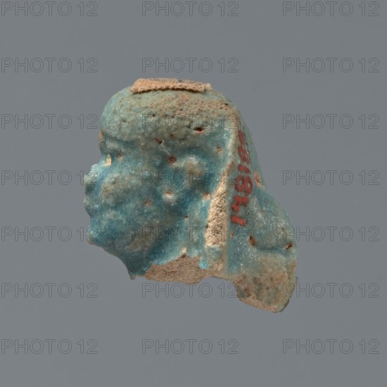 Shabti head, Mummy-shaped figurine head depicting a king. Oval face with full cheeks; prominent eyebrow arch. Eye and nose shapes are difficult to distinguish. Corners of the mouth slightly upturned. Low-set nemes headdress with its edges slightly outlined against the forehead. End of the rear pillar visible at the back of the head. Ptolemaic Period, 332–30 BC, Egypt. Shabti; height 3 cm, width 2.3 cm., A small three-dimensional fragment shaped like a human head in profile, showing a face with a forehead, brow, nose, lips, and chin, rendered with a textured, pitted surface; the fragment has a worn, uneven underside and a flat area on top. The visible colors include turquoise-blue across most of the surface, pale beige in exposed areas and the underside, small patches of brownish spots where the surface is eroded, and a small area of reddish color on one side. The background is a solid neutral gray.
