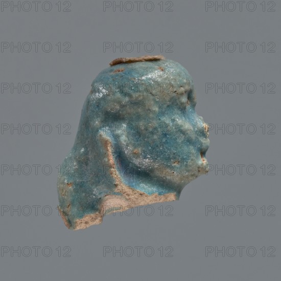 Shabti head, Mummy-shaped figurine head depicting a king. Oval face with full cheeks, prominent eyebrow arch; eye and nose shapes are indistinct. Corners of the mouth slightly upturned. Wears a low-set nemes whose edges are slightly outlined against the forehead. End of the rear pillar visible at the back of the head. Ptolemaic Period, 332–30 BC, Egypt. Shabti; height 3 cm, width 2.3 cm., A small sculpted fragment resembling a head in profile, with a weathered, glossy surface showing turquoise blue, teal, and pale blue-green tones mixed with areas of tan and beige where the material is exposed; the form has softened facial contours, a rounded top with a thin brown strip along the crown, and uneven, eroded edges revealing a rough, sandy interior against a uniform gray background.