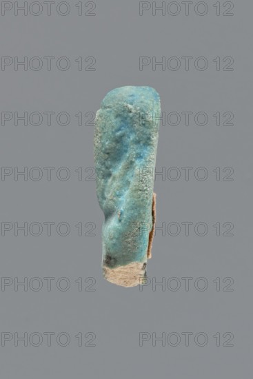 Shabti, Mummy-shaped figurine, funerary gift. Legs missing. Arms crossed on the chest, hands invisible from under the shroud. Long, smooth, three-part wig; wide, straight, smooth beard. Not inscribed. Late Period, Egypt, 664–332 BC. Height 3.8 cm., A vertically oriented, irregularly shaped object with a rough, grainy surface in shades of turquoise, blue, and green against a flat gray background; the object shows areas of lighter sandy beige at its lower edge and a small strip of beige material along one side, with subtle darker blue-green patches and textured, pitted surfaces throughout.