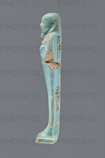 Ushabti, Mummy-shaped figurine, standing on a base with a narrow, slightly convex pillar at the back. Arms crossed high on the chest, right folded over left. Left hand holds a pickaxe, right a mattock; a twisted rope passes over the left shoulder with a bag at the end on the back. Wears a smooth, three-part wig and a long straight beard. Facial details poorly legible. Not inscribed. Ptolemaic Period, 332–30 BC, Egypt. Height 12.1 cm., A narrow turquoise-green figurine in profile against a gray background, showing a stylized human head with a rounded skull and a short beard, a torso with folded arms, and an elongated lower body tapering to a flat foot; the surface has areas of lighter beige where the material is worn or chipped and spots of darker blue-green patina.