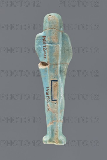Ushabti, Mummy-shaped figurine, standing on a base with a narrow, slightly convex pillar at the back. Arms crossed high on the chest, right folded over left. Left hand holds a pickaxe, right a mattock; a twisted rope passes over the left shoulder with a bag at its end on the back. Wears a smooth, three-part wig and a long straight beard. Facial details poorly legible. Not inscribed. 332–30 BC, Ptolemaic Period, Egypt. Height 12.1 cm., A small human-shaped figurine viewed from the back, made of a smooth turquoise-blue material with areas of beige where the surface is worn or chipped; the figure has a rounded head, broad shoulders, a long narrow body, and indistinct feet. The surface shows a few cracks and worn patches and a small hollowed area on one side; there is a rectangular inset on the back with a thin dark-blue border. The background is a uniform mid-gray.