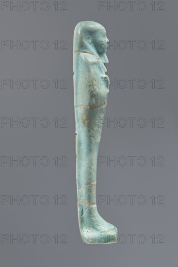 Ushabti, Mummy-shaped figurine, funerary offering, standing on a base with a narrow, slightly convex pillar at the back. Arms crossed high on the chest, right folded over left. Left hand holds a pickaxe, right a mattock; a twisted rope passes over the left shoulder with a bag on the back end. Smooth, three-part wig, long straight beard; facial details poorly legible. Not inscribed. Ptolemaic Period, 332–30 BC, Egypt. Height 12.1 cm., A narrow vertical statuette of a standing human figure shown in profile against a plain gray background, with a smooth elongated form and a rounded head wearing a close-fitting headdress; the figure’s arms are folded across the chest and the legs are together, ending in a flat, slightly rounded foot on a short base. The surface shows a worn, weathered texture with small pits and abrasions and visible joins where sections meet. The dominant color of the object is a turquoise-blue with areas of lighter blue-green and some pale beige or tan patches along the ...
