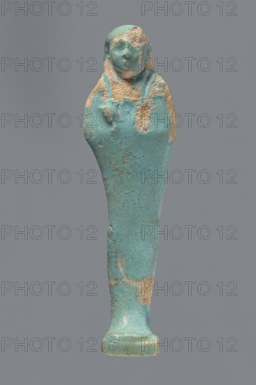 Ushabti. Mummy-shaped figurine standing on a base with a flat pillar at the back. Arms folded across the chest with delicately outlined hands visible from under the shroud; both hands hold hoes and a basket is slung over the left shoulder on the back. Wears a smooth, tripartite wig. Round, proportionate face. Not inscribed. 664–332 BC, Late Period, Egypt. Height 11 cm., A single small statuette shown against a plain gray background, with a turquoise-blue surface mottled with areas of beige where the coating has worn away; the figure has a rounded head with faint facial features and a suggestion of a headdress, a tapered body that widens slightly at the shoulders, a narrow lower section leading to a small flared base, and an overall textured, aged appearance with patches of turquoise and beige.