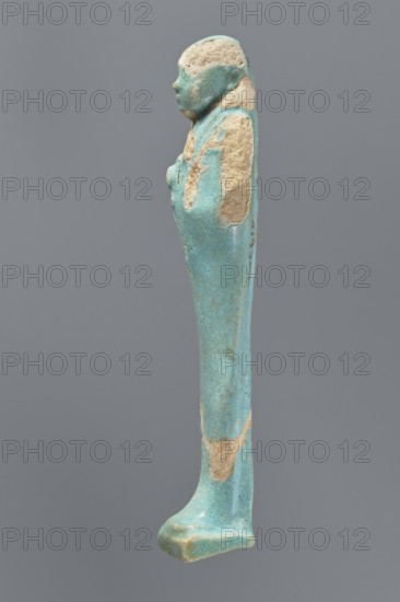 Ushabti, mummy-shaped figurine. Standing on a base with a flat pillar at the back. Arms folded across the chest, with delicately outlined hands visible from under the shroud. Both hands hold hoes, and a basket is slung over the left shoulder on the back. Smooth tripartite wig. Round, proportionate face. Not inscribed. 664–332 BC, Late Period, Egypt. Height 11 cm., A slender figurine in shades of turquoise with areas of beige wear, shown in profile against a plain gray background, depicting a standing humanlike form with a rounded head, a suggested facial profile, a long narrow body, folded arms against the chest, and a flared base forming the feet; the surface shows smooth glazed turquoise areas interrupted by rough beige patches and slight darker blue-green variations.