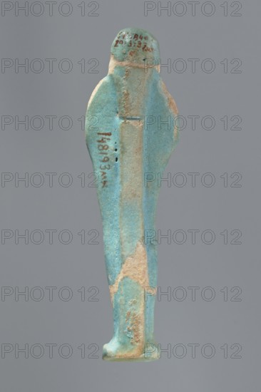 Ushabti, Mummy-shaped figurine standing on a base with a flat pillar at the back. Arms folded across the chest with delicately outlined hands visible from under the shroud; both hands hold hoes and a basket is slung over the left shoulder on the back. Wearing a smooth tripartite wig; round face with proportionate facial details. Not inscribed. 664–332 BC, Late Period, Egypt. Height: 11 cm., A slender figurine viewed from the back, primarily turquoise blue with areas of pale beige where the surface is worn, showing a narrow vertical ridge along the center back, rounded shoulders, a small rounded head, and a rectangular base; the surface has a slightly rough texture with some small pits and patches of darker blue and tan.