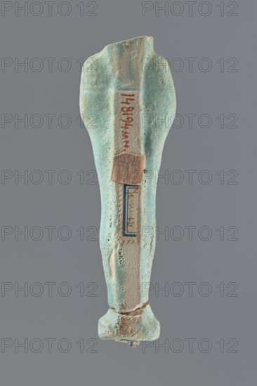 Shabti, Mummy-shaped figurine, funerary offering, standing on a base; head missing. A narrow, slightly convex pillar at the back. Arms crossed high on the chest, right folded over left. Left hand holds a pickaxe, right a mattock, with a twisted rope passed over the left shoulder; a vertically hanging bag at the end of the rope on the back. Convex tools; handles of both tools slightly curled at the ends. Smooth tripartite wig and short, straight beard. Not inscribed. Late Period, Egypt, 664–332 BC. Shabti; height 11 cm., A single elongated object with a rounded top and a wider base, shown against a neutral gray background, appears worn and pitted with an uneven surface texture. The object is predominantly turquoise and light blue with areas of pale beige and off-white where the surface is more eroded. A small rectangular patch of tan fabric is attached near the upper middle, and a narrow band of darker blue framing is visible beneath that patch. There are spots of brownish discoloration...