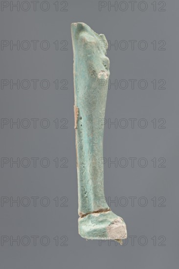 Shabti, Mummy-shaped figurine, standing on a base; head missing. A narrow, slightly convex pillar at the back. Arms crossed high on the chest, right folded over left. Left hand holds a pickaxe, right a mattock; a twisted rope passes over the left shoulder, ending on the back in a vertically hanging bag. Convex tools with handles slightly curled at the ends. Smooth tripartite wig and short straight beard. Not inscribed. 664–332 BC, Late Period, Egypt. Shabti; height 11 cm., A vertical elongated object with a weathered, rough surface in shades of turquoise and light green, with areas of pale beige and off-white where the outer layer is chipped, set against a solid medium-gray background; the object tapers slightly toward the top, shows pitted texture and irregular edges, and rests on a thicker base portion with a broken, uneven bottom.
