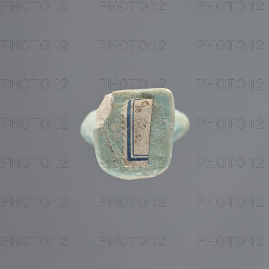 Shabti, Mummy-shaped figurine, standing on a base; head missing. A narrow, slightly convex pillar at the back. Arms crossed high on the chest, right folded over left. In the left hand a pickaxe, in the right a mattock, and a twisted rope passed over the left shoulder; on the back at the end of the rope a vertically hanging bag. Convex tools with handles slightly curled at the ends. Smooth, tripartite wig and short, straight beard. Not inscribed. 664–332 BC, Late Period, Egypt. Height: 11 cm., A small rounded object with a weathered, uneven surface in shades of pale turquoise and light blue sits against a neutral gray background, showing a flat top area with rough beige and off-white patches and a thin vertical band of darker blue along one edge; the surface has visible texture and small cracks, and a faint suggestion of a torn paper fragment in muted cream is adhered to the top.