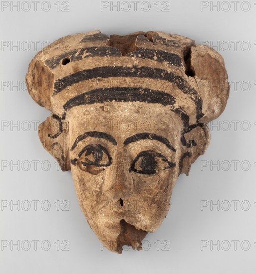 Sarcophagus mask with an elongated, triangular face. A fragment of a wig is preserved above the forehead; the wig is painted with black stripes. Disproportionately large eyes placed asymmetrically in relation to each other. The outline of the eyes, pupils, eyebrows, and details of the ears are highlighted with black dye. 1st–3rd century, Roman Period, Egypt. Sarcophagus mask; height 24.8 cm, width 22.5 cm., A worn carved face mask with a beige-brown surface and dark black painted accents, showing a rounded top with a striped band of dark black arcs, stylized eyebrows and eye outlines in black, hollowed eye openings with dark interior areas, a carved nose with a small hole, and a damaged lower chin with missing fragments; the ears are visible at the sides and the overall texture is rough and aged, with variations of beige, brown, and black across the piece.