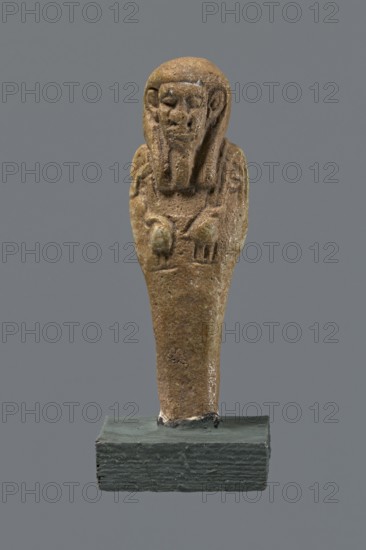 Shabti, mummy-shaped figurine standing on a base, back supported by a pillar/pilaster. Arms crossed on the chest with only the hands visible from under the shroud; right hand holds a hoe and left hand a pickaxe. A basket is slung on a string over the left shoulder on the back. Wears a three-part smooth wig and a long straight smooth beard. Features large eyes, a wide nose, a grimacing mouth, and very large ears. Inscribed inscription on the pilaster illegible. 332–30 BC; Late Period–Ptolemaic period, Egypt. Shabti; height 8.5 cm., A small sculpted figure of brownish stone with a rounded head and a stylized face wearing a headdress, the arms crossed over the chest and the lower body tapering into a narrow base, mounted on a dark greenish-gray rectangular stand against a solid medium-gray background; visible colors include shades of brown, gray, greenish-gray, and hints of lighter beige.