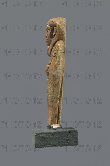 Shabti, Mummy-shaped figurine, funerary gift, standing on a base with back supported by a pillar/pilaster. Arms crossed on the chest with only the hands visible from under the shroud; right hand holds a hoe and left a pickaxe. A basket is slung on a string over the left shoulder on the back. Wears a three-part smooth wig; beard long, straight, and smooth. Large eyes, wide nose, grimacing mouth, very large ears. Inscribed inscription on the pilaster illegible. 332–30 BC; Late Period–Ptolemaic period, Egypt. Shabti; height 8.5 cm., A slender vertical carved figure in shades of brown and tan stands on a rectangular dark grayish base, shown in profile against a uniform gray background; the figure has a rounded head, facial features and a long draped form with subtle texture and weathered surface variations in beige and light brown, and the base has a rough, slightly pitted texture in dark gray with a small lighter patch where the figure meets the base.