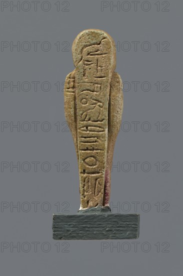 Shabti, mummy-shaped figurine, funerary gift. Mummy-shaped figurine standing on a base, back supported by a pillar/pilaster. Arms crossed on the chest, only the hands visible from under the shroud; right hand holds a hoe, left hand a pickaxe. On the back is a basket slung on a string over the left shoulder. Wearing a three-part smooth wig; long, straight, smooth beard. Large eyes, wide nose, grimacing mouth, very large ears. Inscribed inscription on the pilaster illegible. 332–30 BC; Late Period–Ptolemaic period, Egypt. Shabti, height 8.5 cm., A vertical carved figurine with a rounded top and tapered body in earthy tan and greenish hues standing on a small rectangular dark gray base, showing a textured surface with small pits and a few darker brown spots, and a narrow strip of muted red along one side, set against a uniform medium-gray background.