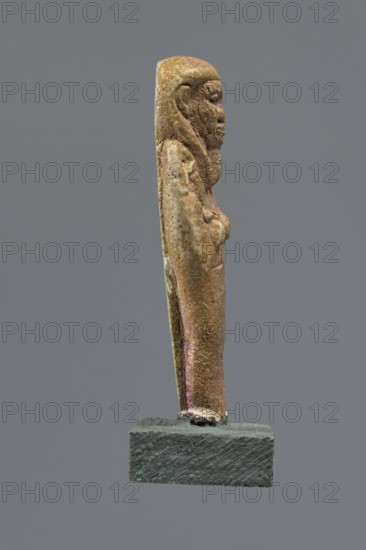 Shabti, Mummy-shaped figurine standing on a base, back supported by a pillar/pilaster. Arms crossed on the chest with only the hands visible from under the shroud; right hand holds a hoe and left hand holds a pickaxe. A basket is slung on a string over the left shoulder on the back. Wears a three-part smooth wig and a long, straight, smooth beard. Large eyes, wide nose, grimacing mouth, very large ears. Inscription placed on a pilaster — illegible. 332–30 BC; Late Period–Ptolemaic period, Egypt. Shabti, height 8.5 cm., A slender vertical statue of a standing figure shown in profile on a small rectangular base, with a textured surface and worn details; the figure has a defined head with facial features, a rounded chest, and arms held close to the body, and the back shows a shallow groove running down its length; visible colors include tan, beige, brown, hints of green and faint pink on the lower side, and the base is a dark grayish color against a solid mid-gray background.