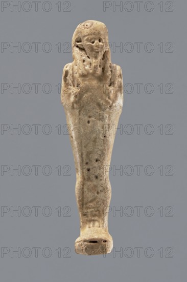Shabti, Mummy-shaped figurine, funerary gift, standing on a base, back supported by a pillar/pilaster. Arms crossed on the chest, right folded over the left. A pickaxe in the left hand, a hoe in the right. On the back is a basket, slung by a single string over the left shoulder. On the head is a three-part, smooth wig. The beard is long, smooth, and straight. The face has well-proportioned features, the mouth set in a slight smile. Painted inscription on the front of the legs in a single wide column framed with lines illegible. Late Period, Egypt, 664–332 BC. Shabti; height 11.1 cm., A small carved figurine in beige and light brown tones stands upright against a solid gray background, showing a stylized human form with a rounded head, faint facial features, and long hair or headdress that falls to the shoulders; the body tapers down into a narrow wrapped or draped lower form with subtle surface pitting and darker brown speckling, and a flat base at the bottom with a few small indentati...