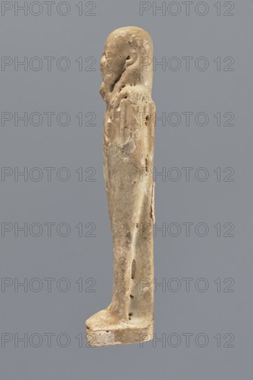 Shabti, Mummy-shaped figurine, standing on a base, back supported by a pillar/pilaster. Arms crossed on the chest, right folded over the left. A pickaxe in the left hand, a hoe in the right. On the back is a basket slung by a single string over the left shoulder. Wearing a three-part smooth wig. Beard long, smooth, and straight. Face with well-proportioned features and a slight smile. Painted inscription on the front of the legs in a single wide column framed with lines; text illegible. 664–332 BC, Late Period, Egypt. Height 11.1 cm., A weathered beige statue of a standing human figure shown in profile against a solid gray background, with worn surfaces, pitted texture, a rounded head, indistinct facial features, a long narrow body and arms close to the sides, and a rectangular base; visible colors include beige, off-white, light brown, and gray.