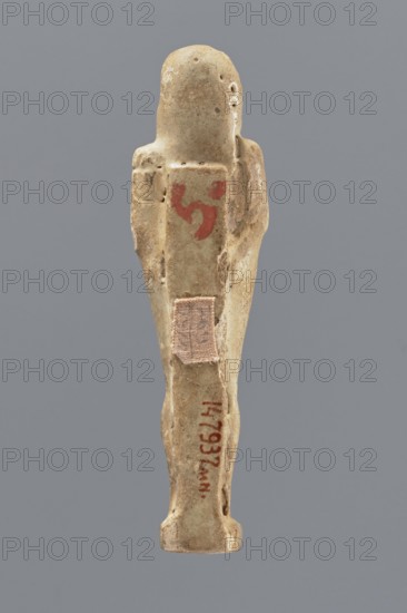 Shabti, Mummy-shaped figurine, standing on a base, back supported by a pillar/pilaster. Arms crossed on the chest, right folded over the left. A pickaxe in the left hand, a hoe in the right. On the back is a basket, slung by a single string over the left shoulder. On the head is a three-part, smooth wig. The beard is long, smooth, and straight. The face has well-proportioned features, the mouth is set in a slight smile. Painted inscription placed on the front of the legs in a single wide column framed with lines; text illegible. 664–332 BC; Late Period, Egypt. Shabti, height 11.1 cm., An elongated small statuette of a person viewed from the back, made of a light beige to tan material with areas of darker brown discoloration and surface wear; the figure has simplified arms alongside the body and a rounded head, with a few small surface pits and rough patches; a small rectangular piece of faded fabric in a pale pinkish-beige tone is attached on the lower back; there is a smudge of red pi...