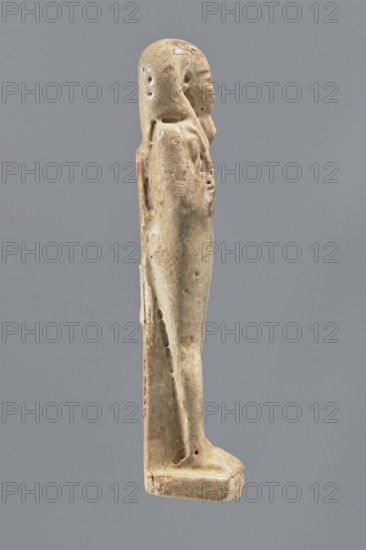Shabti, Mummy-shaped figurine, standing on a base, back supported by a pillar/pilaster. Arms crossed on the chest, right folded over the left. A pickaxe in the left hand, a hoe in the right. A basket on the back, slung by a single string over the left shoulder. Wearing a three-part smooth wig and a long smooth straight beard. Well-proportioned face with a slight smile. Painted inscription on the front of the legs in a single wide column framed with lines; text illegible. Late Period, Egypt, 664–332 BC. Height 11.1 cm., A small carved figure shown in profile against a gray background, rendered in off-white and beige tones with areas of darker brown discoloration and small patches of reddish color; the figure stands on a flat base and has a rounded head, an elongated body, and indications of a cloak or draped garment along the back and sides, with surface pitting and irregularities visible across the material.