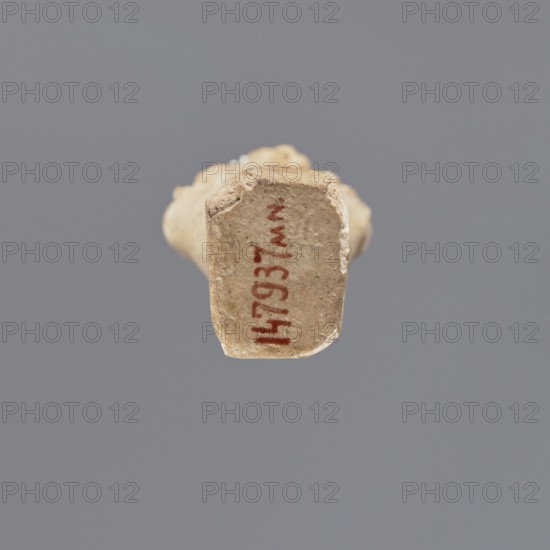 Shabti, Mummy-shaped figurine, funerary gift, standing on a base with back supported by a pillar/pilaster. Arms crossed on the chest, right folded over the left. A pickaxe in the left hand and a hoe in the right. A basket on the back, slung by a single string over the left shoulder. Wearing a three-part smooth wig; long, smooth, straight beard. Well-proportioned face with a slight smile. Painted inscription on the front of the legs in a single wide column framed with lines; text illegible. Late Period, Egypt, 664–332 BC. Height 11.1 cm., A worn beige clay object viewed from one end against a flat gray background, showing a roughly rectangular face with chipped and uneven edges and a few small darker speckles; the object has a vertical streak of reddish-brown color on its face and lighter off-white areas around the edges, with the rest of the image dominated by gray and beige tones.