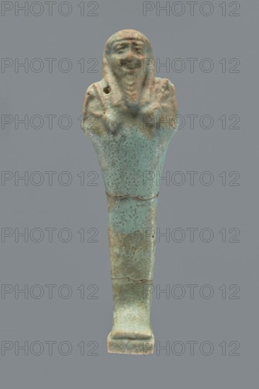 Ushabti, Mummy-shaped figurine shabti, 664–332 BC, Late Period, Egypt. Standing on a base with a flat pillar behind it. Arms folded across the chest with only the palms protruding from under the shroud; a hoe in the right hand and a pickaxe in the left, both tools convex with slightly curled handle ends. A twisted rope is slung over the left shoulder in the right hand, with a trapezoidal basket hanging from the rope on the back. Smooth three-part wig, long braided straight beard, round face with proportionate facial details. Not inscribed. Height 12.2 cm., A small elongated figurine with a rounded top resembling a human head and shoulders, a narrow tapered body and a flat rectangular base, showing a textured surface with pitting and areas of wear; the head and shoulders display simple facial features and a suggestion of raised arms across the chest, with a small hole visible near one shoulder; the object shows mottled colors of pale turquoise, greenish-blue, and patches of brown and be...