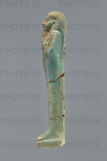 Ushabti, Mummy-shaped figurine shabti. Standing on a base with a flat pillar behind. Arms folded across the chest with only the palms protruding from under the shroud. Holds a hoe in the right hand and a pickaxe in the left; ends of the tool handles slightly curled and tools convex. A twisted rope slung over the left shoulder in the right hand, with a trapezoidal basket hanging from this rope on the back. Smooth three-part wig, long braided beard, round face with proportionate facial details. Not inscribed. Late Period, Egypt, 664–332 BC. Height 12.2 cm., A narrow vertical figurine viewed in profile, predominantly turquoise with areas of greenish-blue and pale blue, showing patches of brownish and tan discoloration and small dark spots; the figure has a rounded head with carved facial features and a long braided or draped hairstyle, a suggestion of crossed arms at the chest, a long straight body that tapers toward the feet, visible horizontal cracks and breaks along the midsection and ...