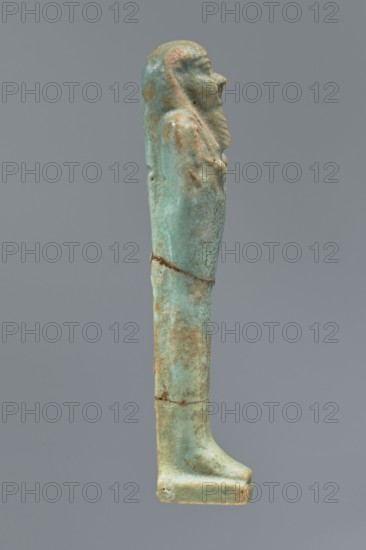 Ushabti, Mummy-shaped figurine shabti. Standing on a base with a flat pillar behind it. Arms folded across the chest with only the palms protruding from under the shroud. A hoe in the right hand and a pickaxe in the left; ends of the handles slightly curled and tools convex. A twisted rope is slung over the left shoulder from the right hand; a trapezoidal basket hangs from this rope on the back. Smooth three-part wig, long straight braided beard. Round, proportionate facial features. Not inscribed. Late Period, Egypt, 664–332 BC. Height 12.2 cm., A slender vertical figurine in shades of green and turquoise with areas of brownish and tan discoloration, shown in profile against a plain gray background; the figure has a rounded head with a pronounced nose and a textured beard, a narrow torso with subtle arm and shoulder contours, a long straight lower body that tapers into a flat rectangular base, and visible surface pitting and weathered patches along its length.