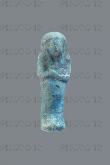 Ushabti, Mummy-shaped figurine impressed in a mold, carelessly made of faience; back flat and unfinished. Arms crossed on the chest, right hand over left; arms distinct. Tools almost invisible — faint outline on the right shoulder suggesting a tool or string for hanging a basket. Smooth three-part wig with front strands reaching to the breasts toward the center of the chest; back strand not cut from the back. Ears exposed, large, vaguely modeled. Facial details illegible; false beard barely visible. Irregularities and lines on the front of the legs suggesting an inscription, now completely illegible. Cracked at waist level and glued together; glaze surface heavily worn., - if there was an inscription, c. 1069–c. 715 BC; Third Intermediate Period, Egypt. Shabti; height 5.6 cm, width 2 cm, depth 1.7 cm., A small blue-green figurine stands upright against a plain gray background; the surface shows a mottled mix of turquoise, cyan, teal, and areas of lighter blue with patches of darker blu...