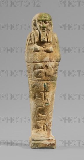 Ushabti, Mummy-shaped funerary figurine, standing on a base and supported at the back by a flat, wide pillar. Arms crossed on the chest with only the hands visible from under the shroud; each hand holds a hoe, the right hand also holds a rope with a basket slung over the left shoulder. Engraved tools. Smooth three-part wig merging at the back into the pillar separated by a thin horizontal line; the rear strand is significantly longer than the front strands. Ears uncovered. Large bulging eyes, wide nose and mouth, long straight beard. Engraved inscription on the front of the figurine's legs. T-shaped; 664–332 BC, Late Period, Egypt. Shabti, height 10.8 cm, width 3 cm., A small sculpted figure with a rounded head and a stylized beard, arms crossed over the chest, and a tapered lower body standing on a rectangular base; the surface is textured with pitting and worn details, showing a sandy beige tone overall with areas of greenish and bluish discoloration and some darker brown spots, set ...